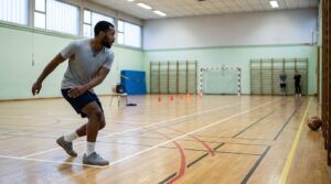 Un jeune homme en tee-shirt gris et short marine pose le pied derrière la ligne blanche pour faire demi-tour pendant un test navette, dans un gymnase municipal au parquet couvert de lignes de terrain, avec espaliers, plots orange et but de handball au fond